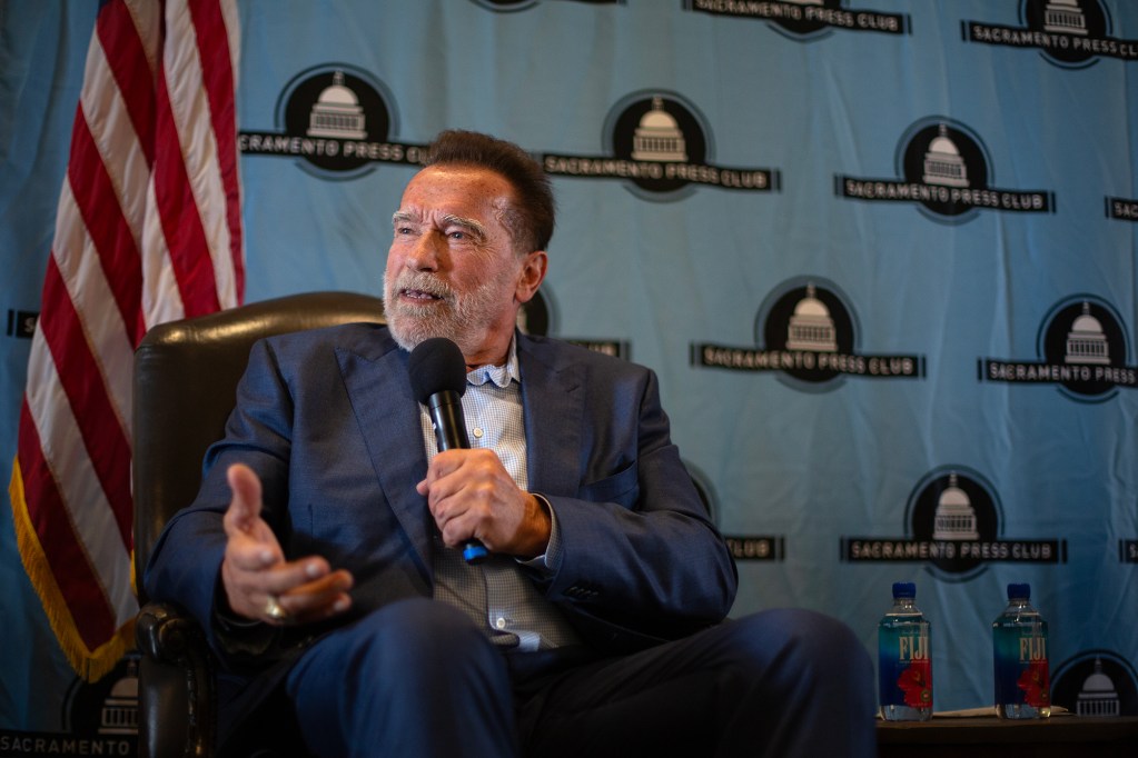 Image of Arnold Schwarzenegger, in blue suit and open collar shirt, looking left and speaking with a microphone in his hand 