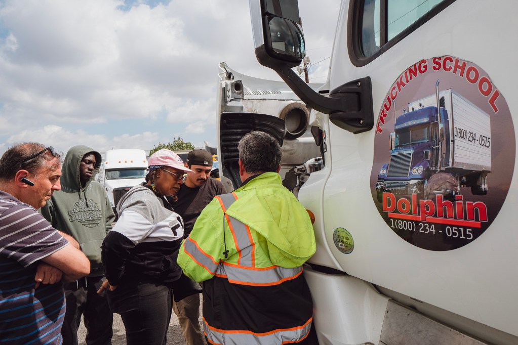 A person dressed in a green and orange safety vest stands next to the engine compartment of a semi-trailer while addressing students as they look-on.