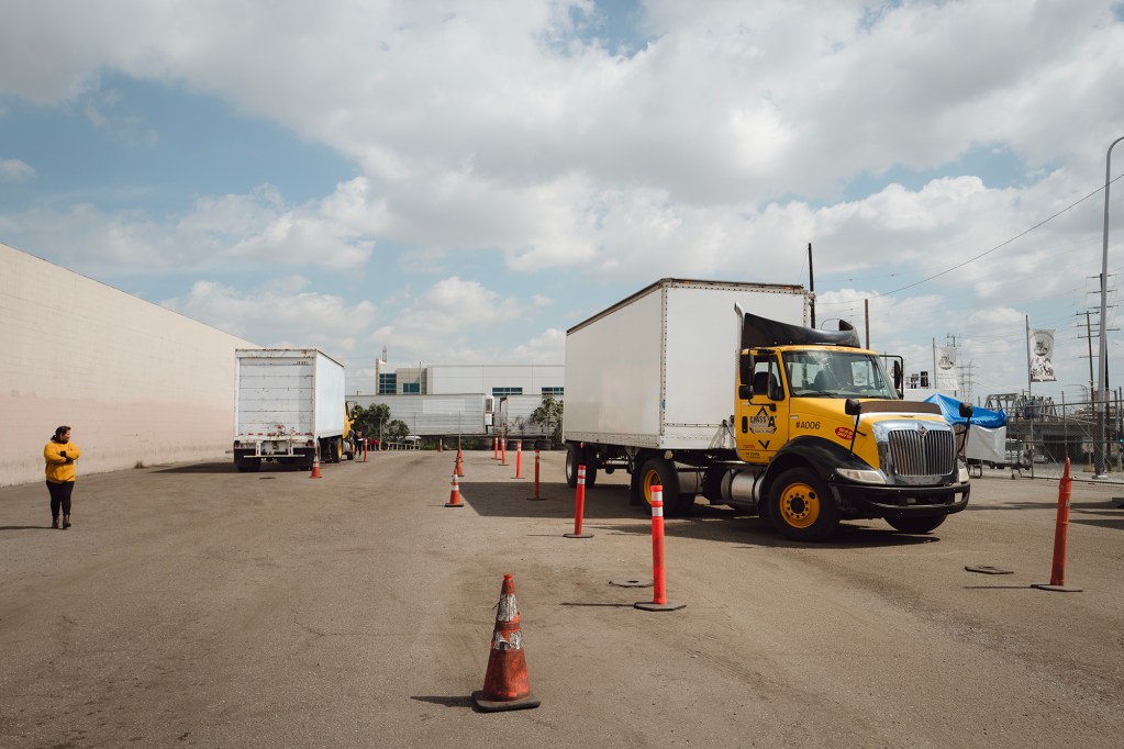 A person wearing black pants and a yellow sweater looks towards a yellow and black semi-trailer pulling a white cargo trailer.
