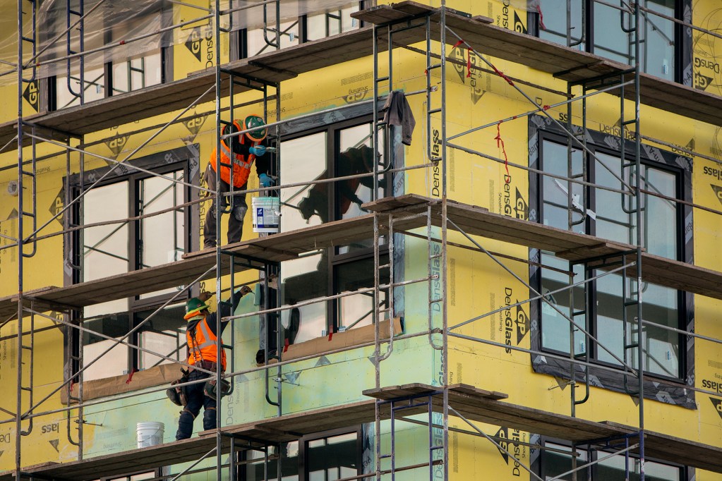 Metal and wood scaffolding frames the corner of a building under construction. Two workers wearing bright orange safety vests and helmets are working on the building. The frame is a close image of the building.