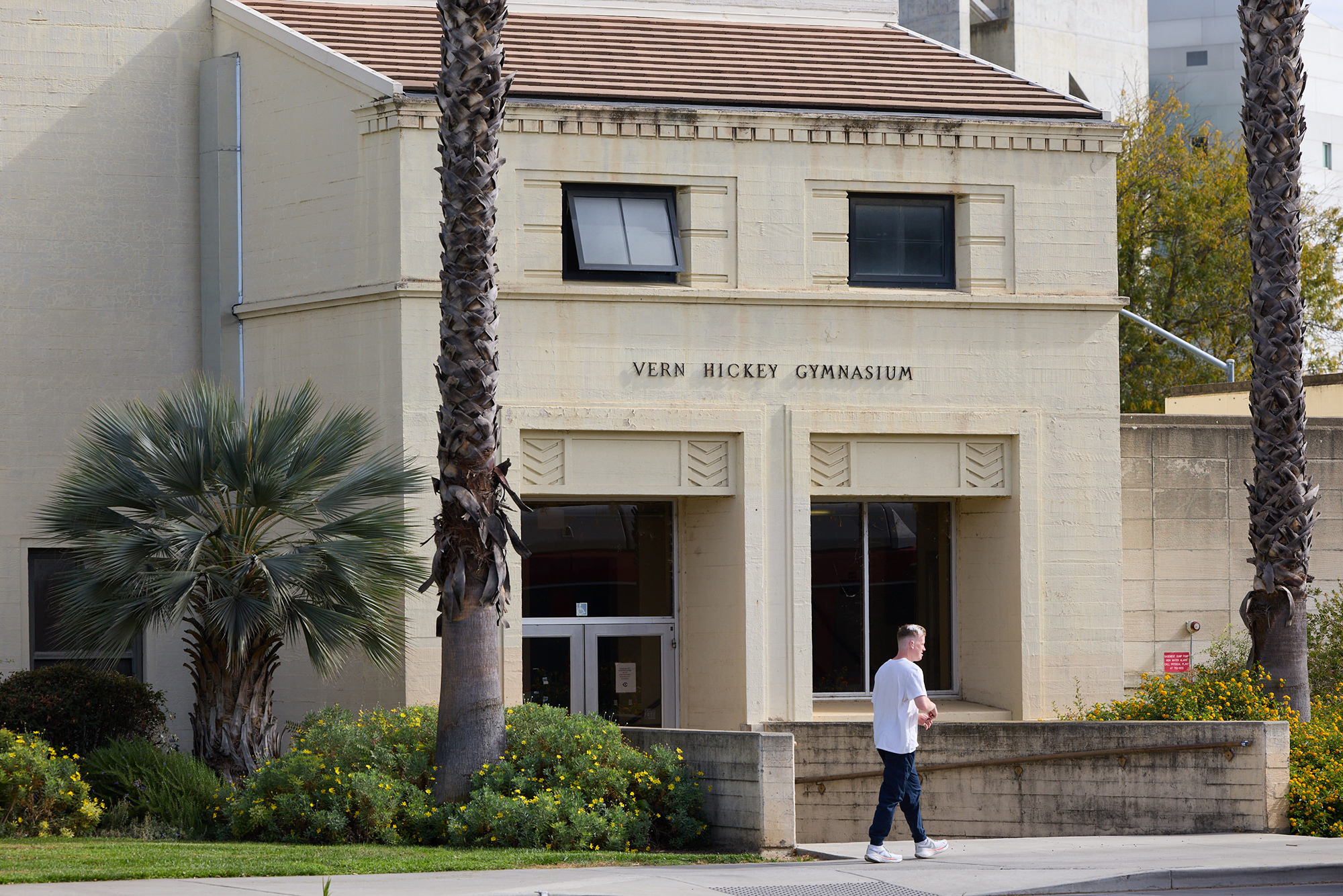 A person walks along the sidewalk in front of an older brick building with two palm trees around the entrance. Over the glass door entrance of the building, it says, “Vern Hickey Gymnasium.”