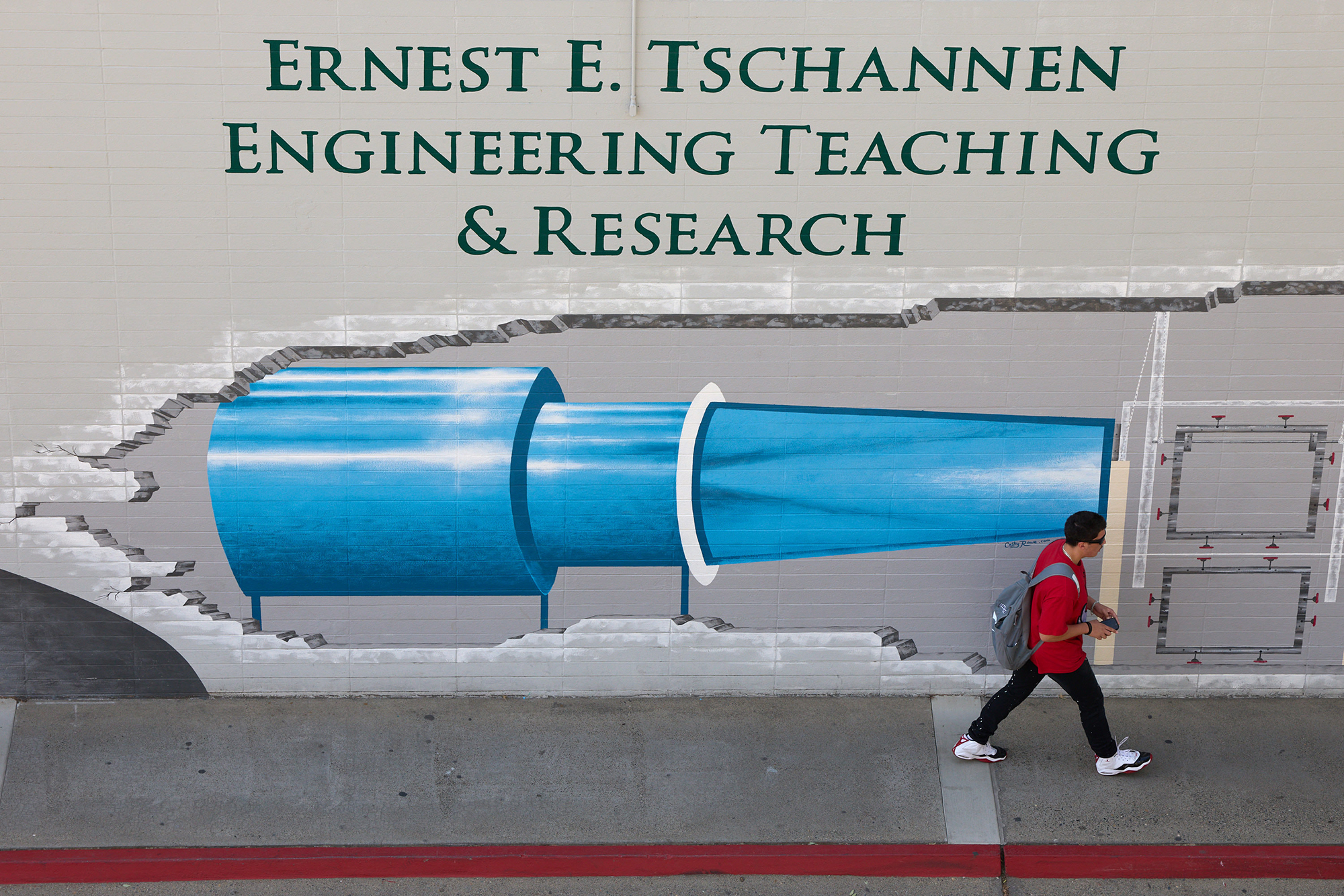 A person walks past a mural of an engine on a grey wall of a college building. The top of the mural says, “Ernest E. Tschannen Engineering Teaching & Research.”