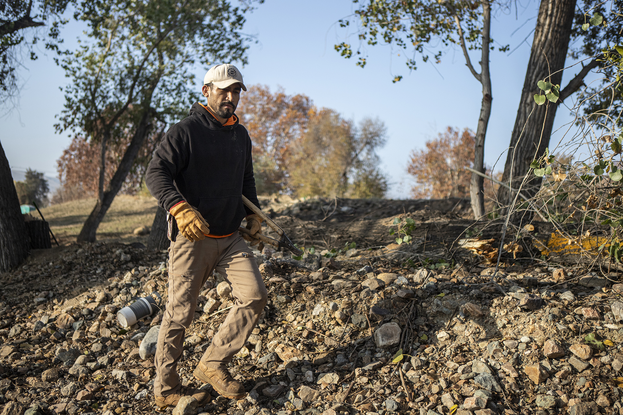 A person in a working sweatshirt and protective work attire walks through rough terrain in a park with pruning shears under their arm.