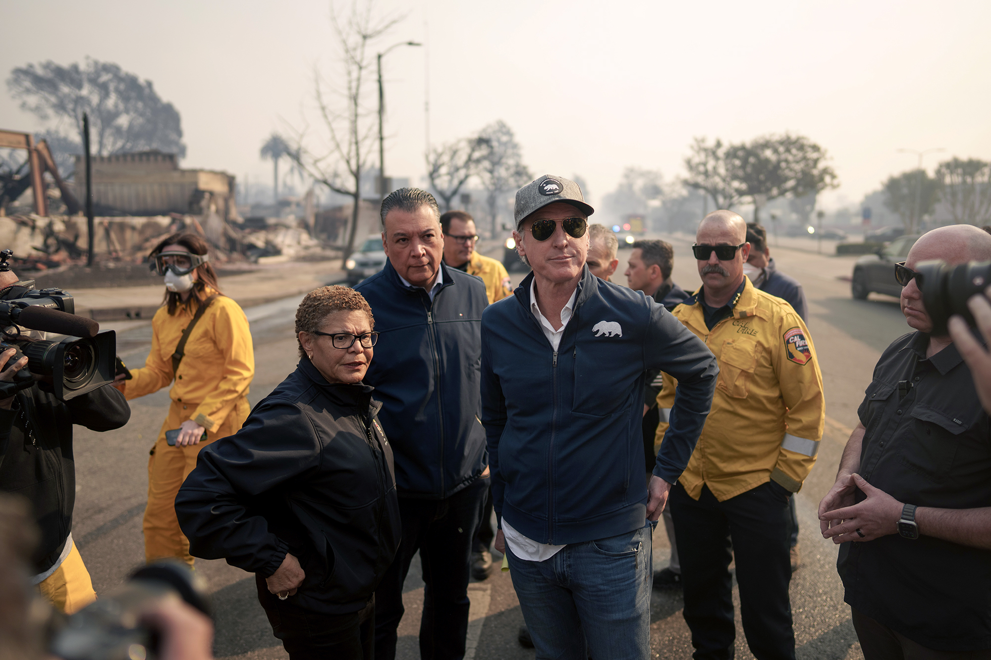 A group of officials and first responders stand on a street in a fire-damaged area, with charred buildings and trees visible in the background. One individual wears a cap and sunglasses, while others wear jackets or uniforms. A camera crew films the scene, and smoke fills the hazy sky.