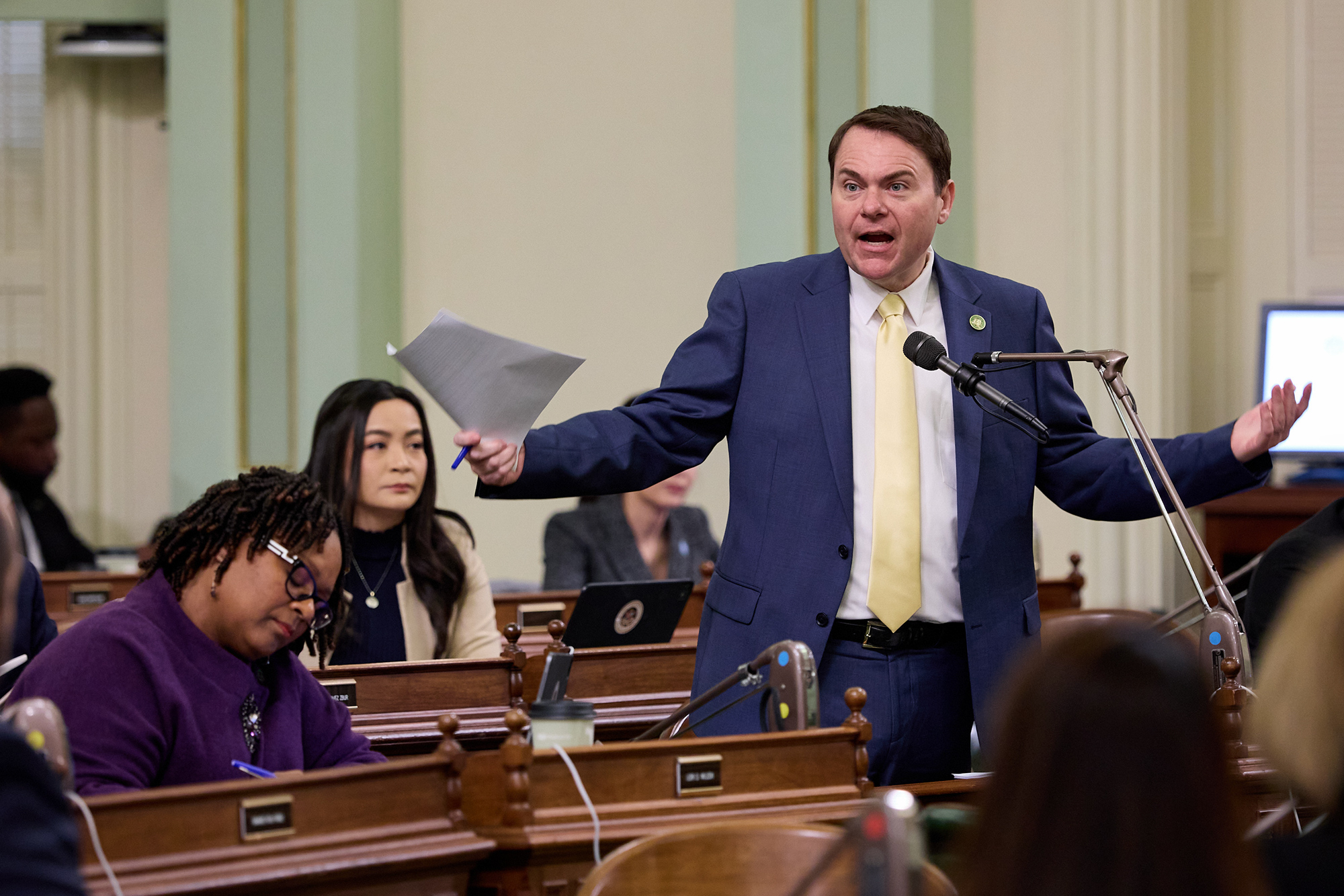 A lawmaker wearing a blue navy suit and bright yellow tie stands in front of a microphone near their desk at the Assembly floor of the state Capitol in Sacramento. The lawmaker extends both their hands out while they hold a piece of paper and pen with in their right hand.