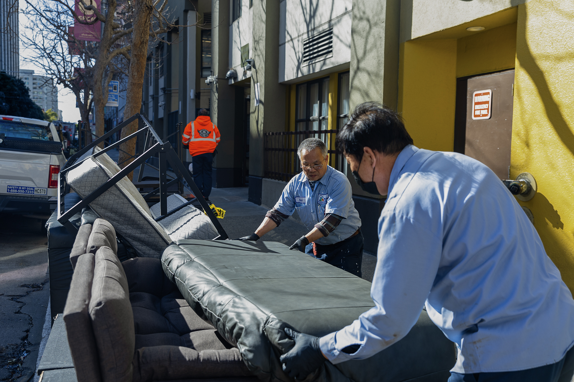 Two workers wearing light blue uniforms and gloves remove a black leather couch from a sidewalk outside a building with yellow and gray walls. Various pieces of discarded furniture, including a gray sofa and metal bed frame, are piled nearby. In the background, a person wearing a bright orange safety jacket walks away from the scene on a city street lined with trees and buildings. Sunlight casts strong shadows on the walls and pavement.