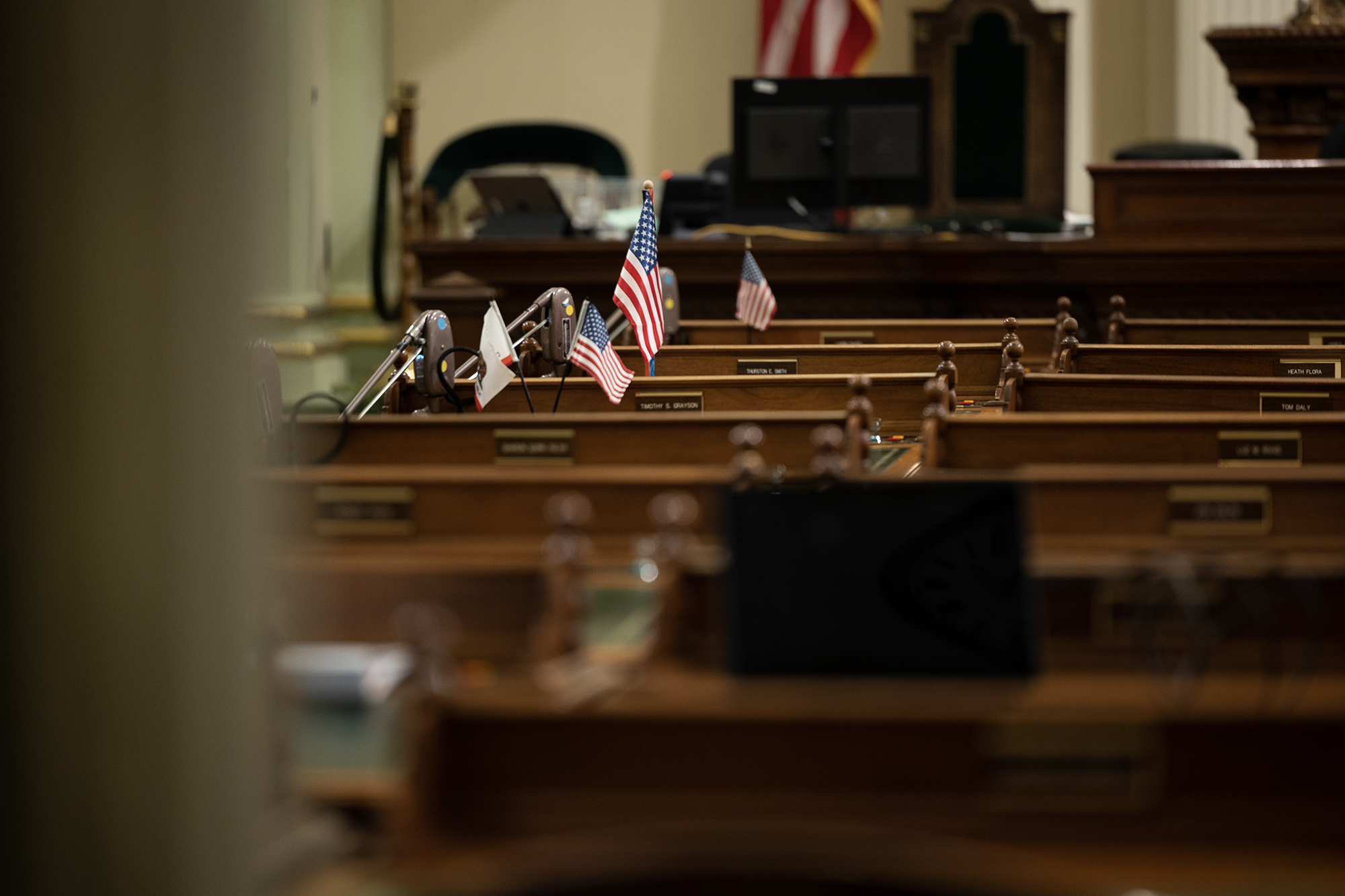 A view of empty desks inside the California State Assembly chamber, with small American and California flags displayed on microphones. Nameplates and monitors are visible in the background.