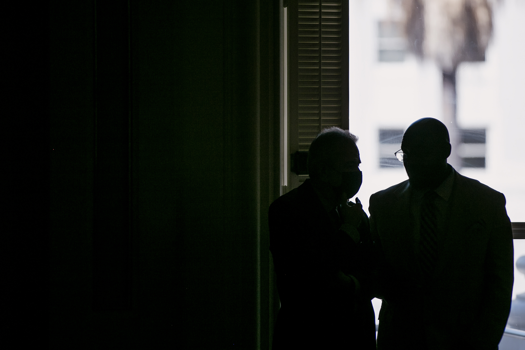 Two silhouetted figures stand in front of a window inside the California Capitol, one wearing a mask, the other wearing glasses and a suit. Light from outside highlights their outlines.