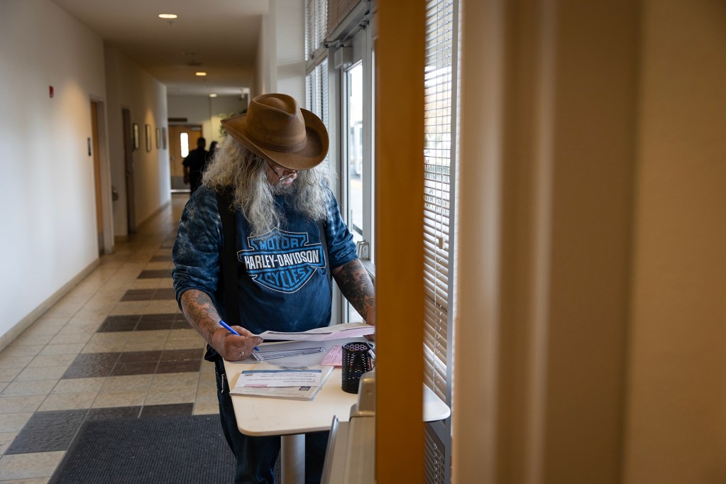 A person wearing a brown cowboy hat, glasses, and a Harley-Davidson t-shirt stands at a small table inside a hallway, filling out paperwork with a pen. Their long, wavy hair and tattoos are visible as they focus on the documents. The setting is a public building with tiled floors, beige walls, and a row of doors leading to other rooms. Natural light streams in through the windows, which have partially closed blinds. A few people are visible in the background walking through the hallway.