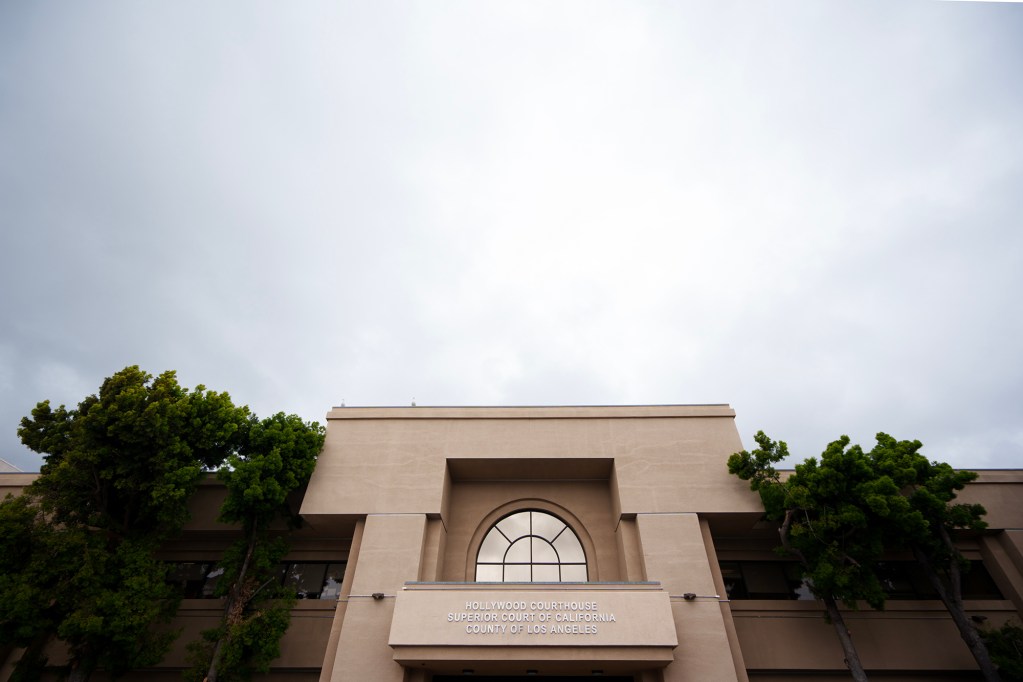 The exterior of the Hollywood Courthouse, a beige concrete building with a large arched window above the entrance. The sign reads “Hollywood Courthouse, Superior Court of California, County of Los Angeles.” Tall trees frame both sides of the building, and the sky above is overcast.