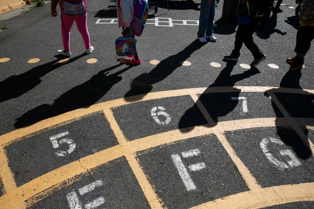 Painted letters and numbers on the asphalt of a school as the feet and shadows of young children can be seen in the background.
