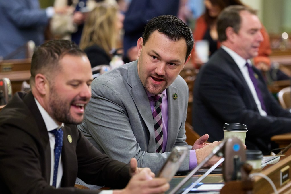 A person looks to their left as they talk with anotehr person while both sit in front of desks at the state Capitol.
