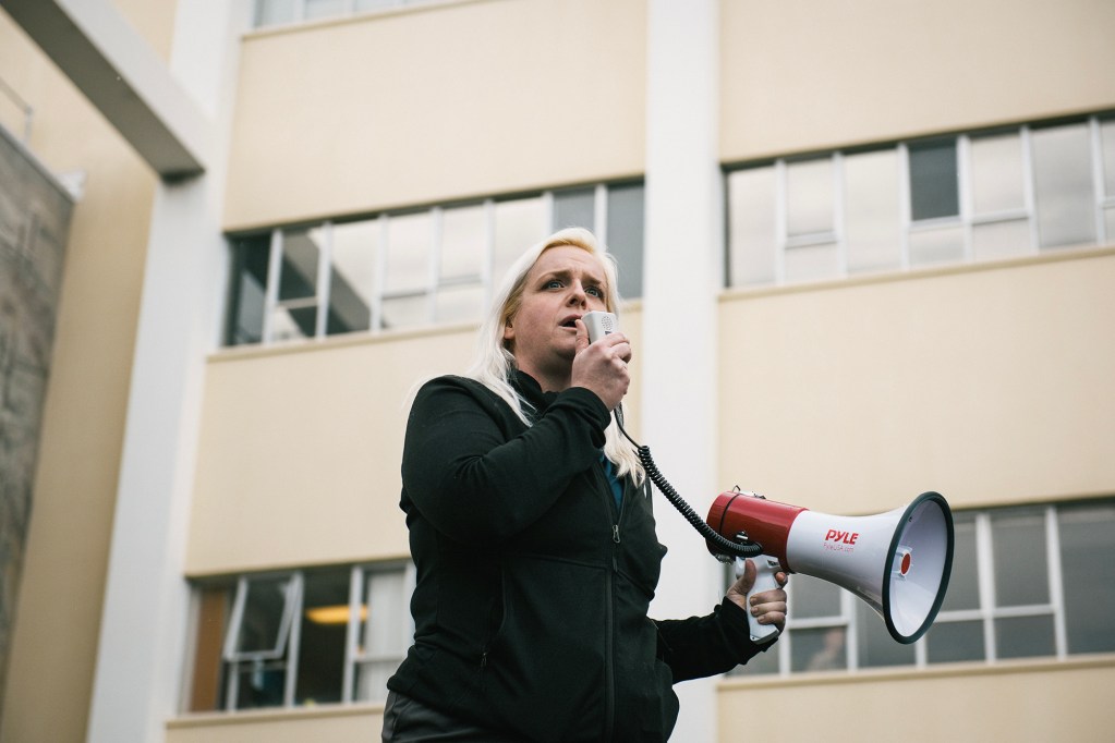 A person with long blond hair speaks into a microphone while holding a red and white megaphone. They are standing outdoors in front of a beige building with multiple windows, appearing to address a crowd. The expression on their face is serious and focused.