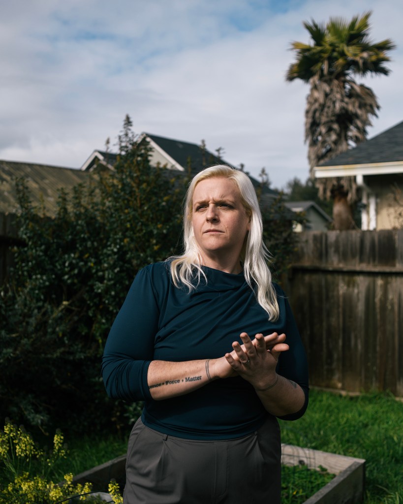A person with long, light blond hair stands in a backyard with a serious expression, looking off into the distance. They wear a navy long-sleeve top and gray pants, with a visible tattoo on their forearm reading “Force = Mass x Acceleration.” Behind them are raised garden beds, a wooden fence, and a palm tree next to a house. The lighting is soft, with clouds and blue sky above.
