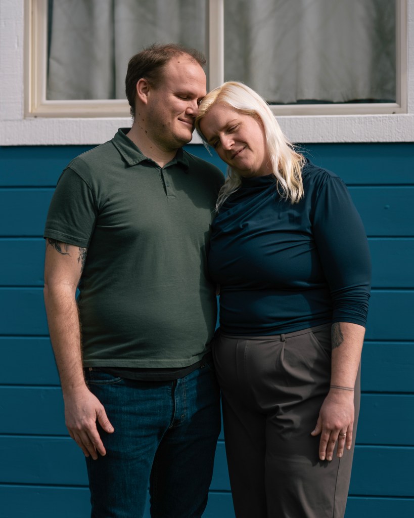 A couple stands closely together in front of a blue and white house. They lean their heads against each other with their eyes closed, sharing a tender moment. The person on the left wears a green polo shirt and jeans, and the person on the right wears a navy top and gray pants.