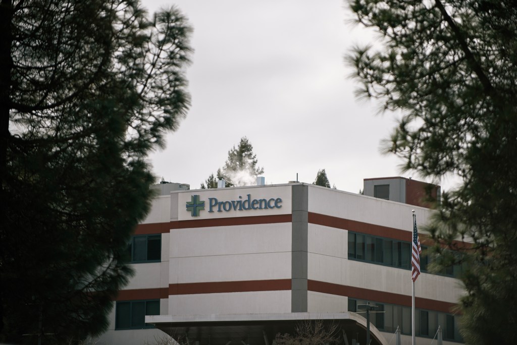 A Providence hospital building framed by pine trees on an overcast day. The sign with the hospital's name and cross logo is clearly visible on the white and red-striped exterior. An American flag flies in front of the entrance.