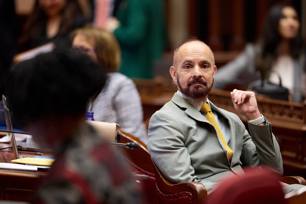 A person wearing a grey blazer with a white shirt and yellow tie looks to their right as they sit in front of a wooden desk surrounded by other people.