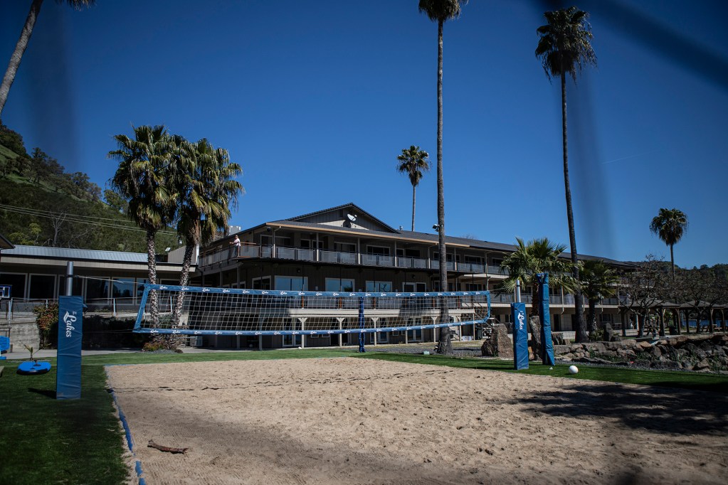 The exterior view of a treatment facility is shown through the net of a volleyball court with sand and grass all around and palm trees scattered around the building on a sunny day.