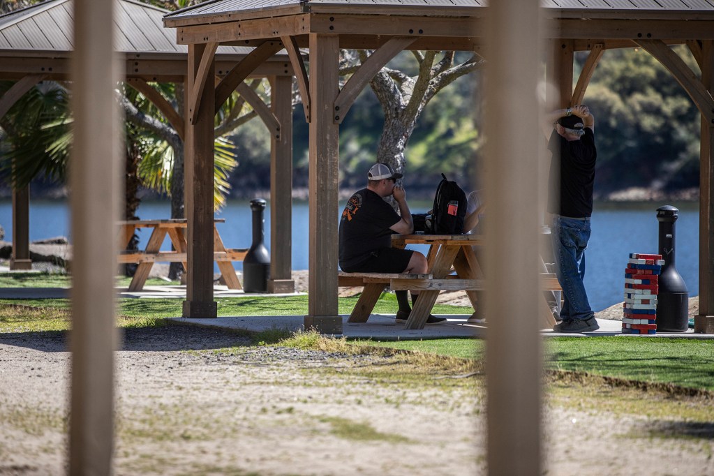 A person wearing a black t-shirt is shown sitting at a picnic table smoking in the shade of a gazebo along a lakeside on a very bright and sunny day.