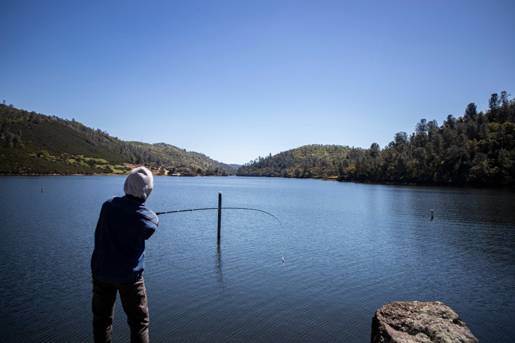 The back view of a person wearing a flannel hoodie with the hood up as they cast their fishing rod into a lake to fish on a sunny day. The background depicts a view of the lake extending to a series of lush green hills.