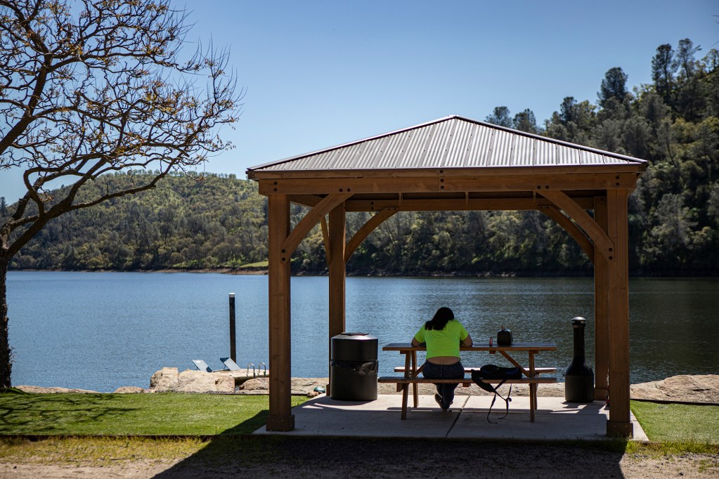A person wearing a yellow t-shirt is shown sitting at a picnic table in the shade of a gazebo along a lakeside on a very bright and sunny day.
