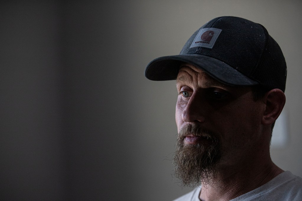 A person with a beard and a baseball cap is shown in a close-up view of their face, displaying a sad expression. Half their face is illuminated from the light coming through a door in a dark room, and a shadow covers the other side of the face.
