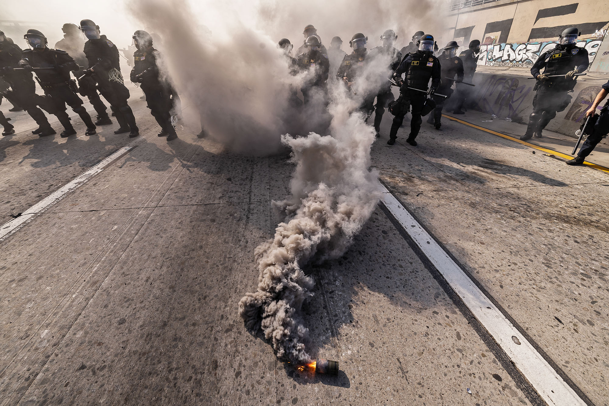 A canister laying in the middle of a freeway road releases fire and clouds of smoke towards law enforcement officers dressed in riot gear during a protest.