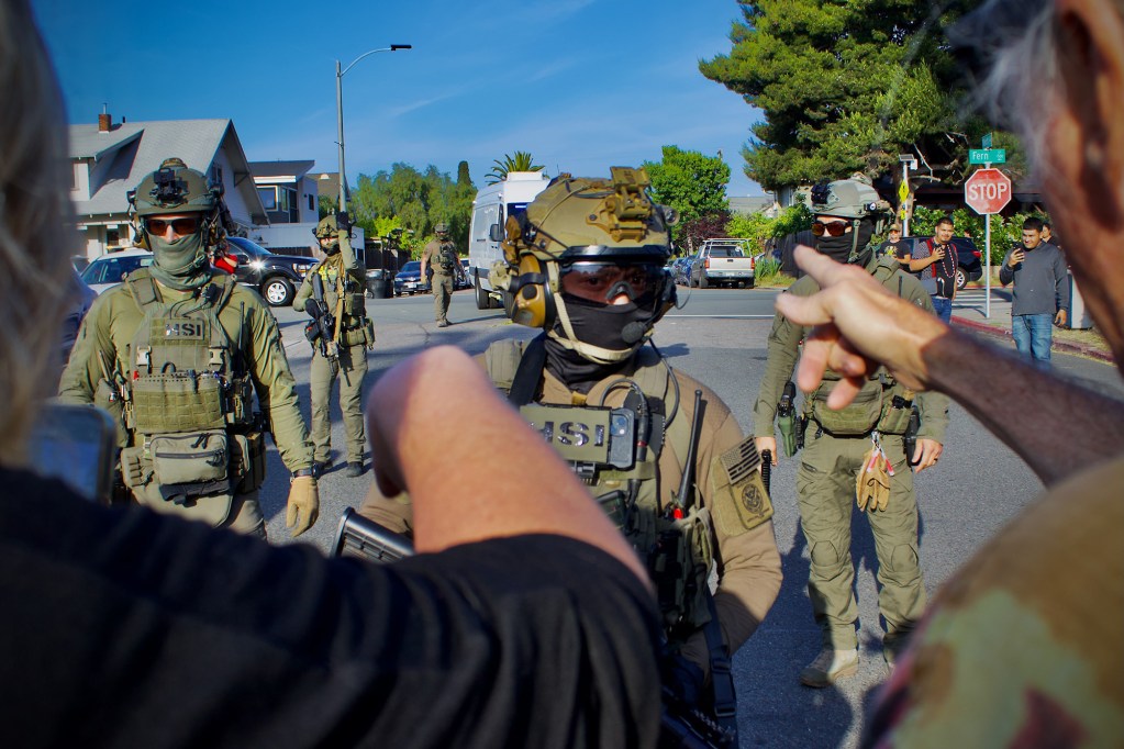 A group of officers, dressed in full tactical gear and wearing face masks, stands in the roadway of a neighborhood street during the middle of the day. In the foreground, we can see over the shoulders of a couple of people as they point to the officers, with one of the officers visible between the two people.
