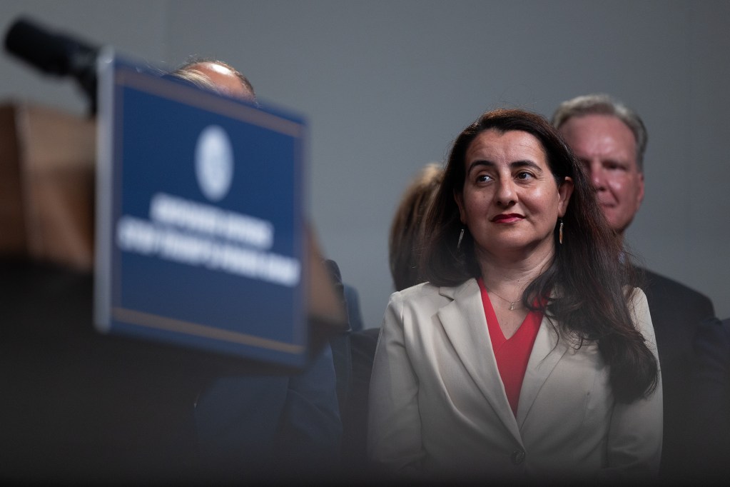A lawmaker, with brown hair and wearing a tan blazer with a red blouse, stands on the side of a lectern and listens to another person speak during a press conference.