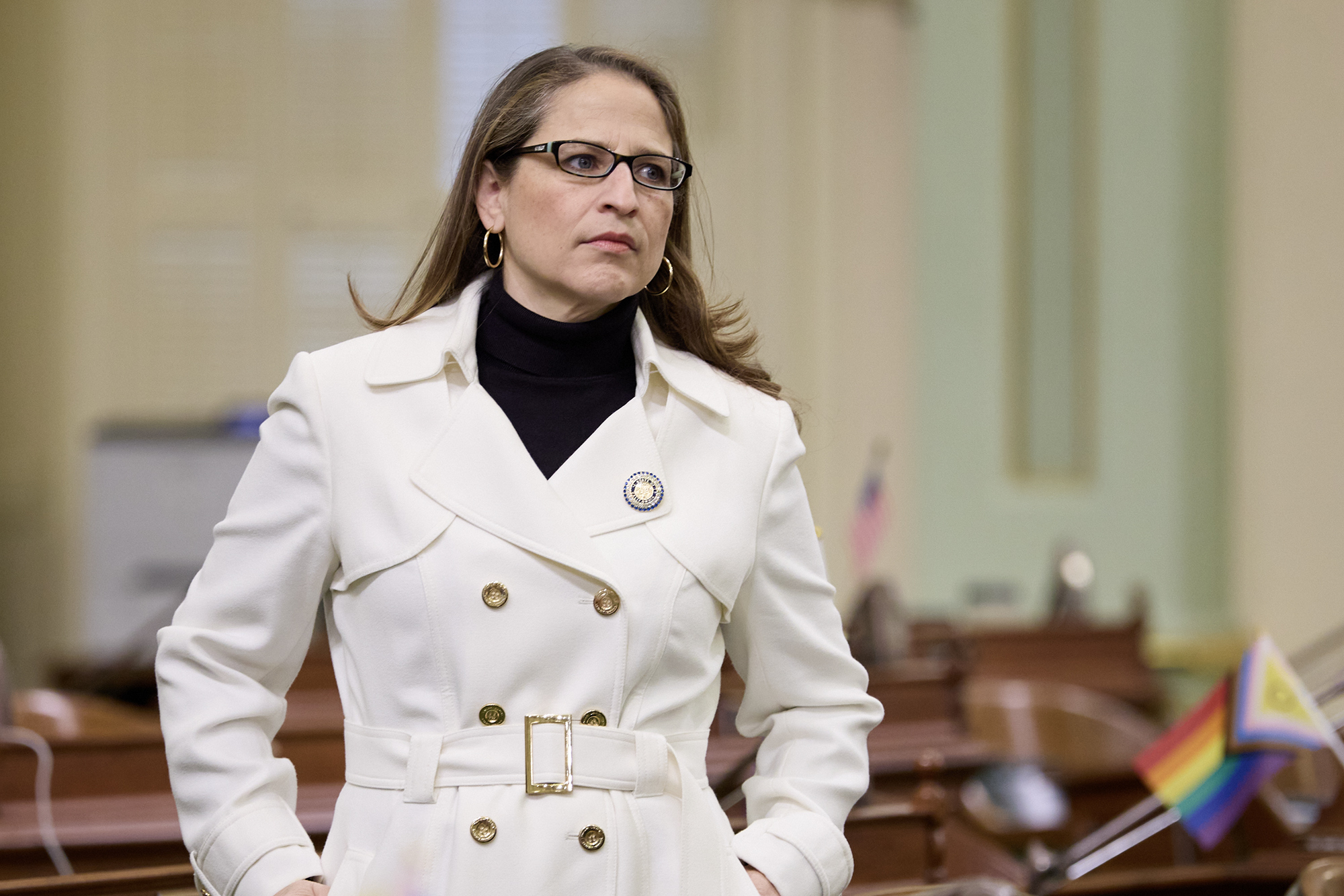 A person wearing glasses and a white trench coat stands inside a government chamber with hands in pockets, looking to the side with a serious expression.
