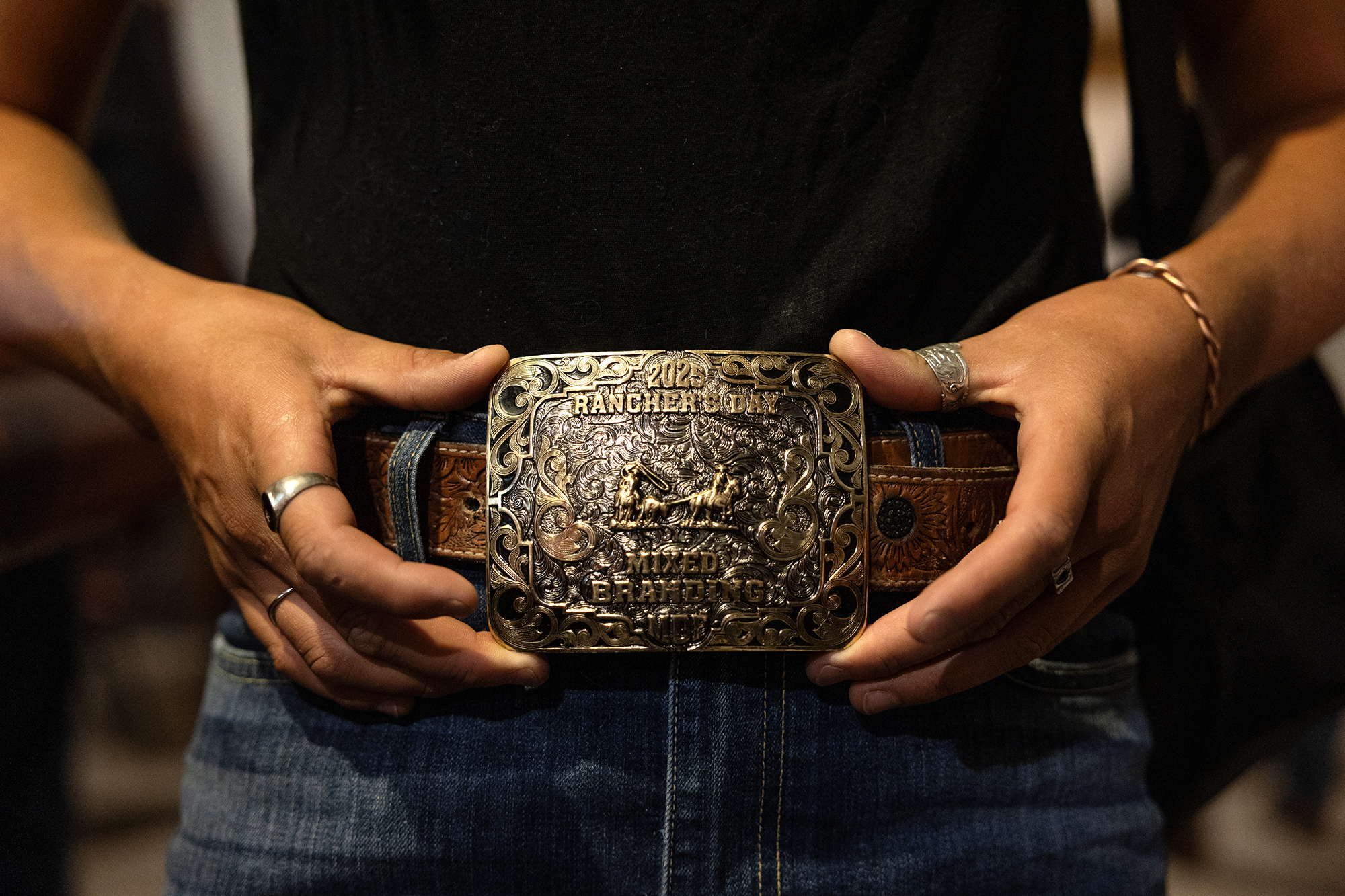 Close-up of a person wearing a large, ornate belt buckle engraved with “2022 Rancher’s Day” and decorative scrollwork, fastened to a tooled leather belt. The person’s hands rest on either side of the buckle, with rings visible on their fingers, and denim jeans are partly visible.