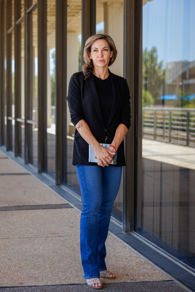 A person stands outside against a glass wall, holding a book with both hands. They are wearing a black blazer, black shirt, blue jeans, and open-toed shoes, with a tattoo visible on one forearm.