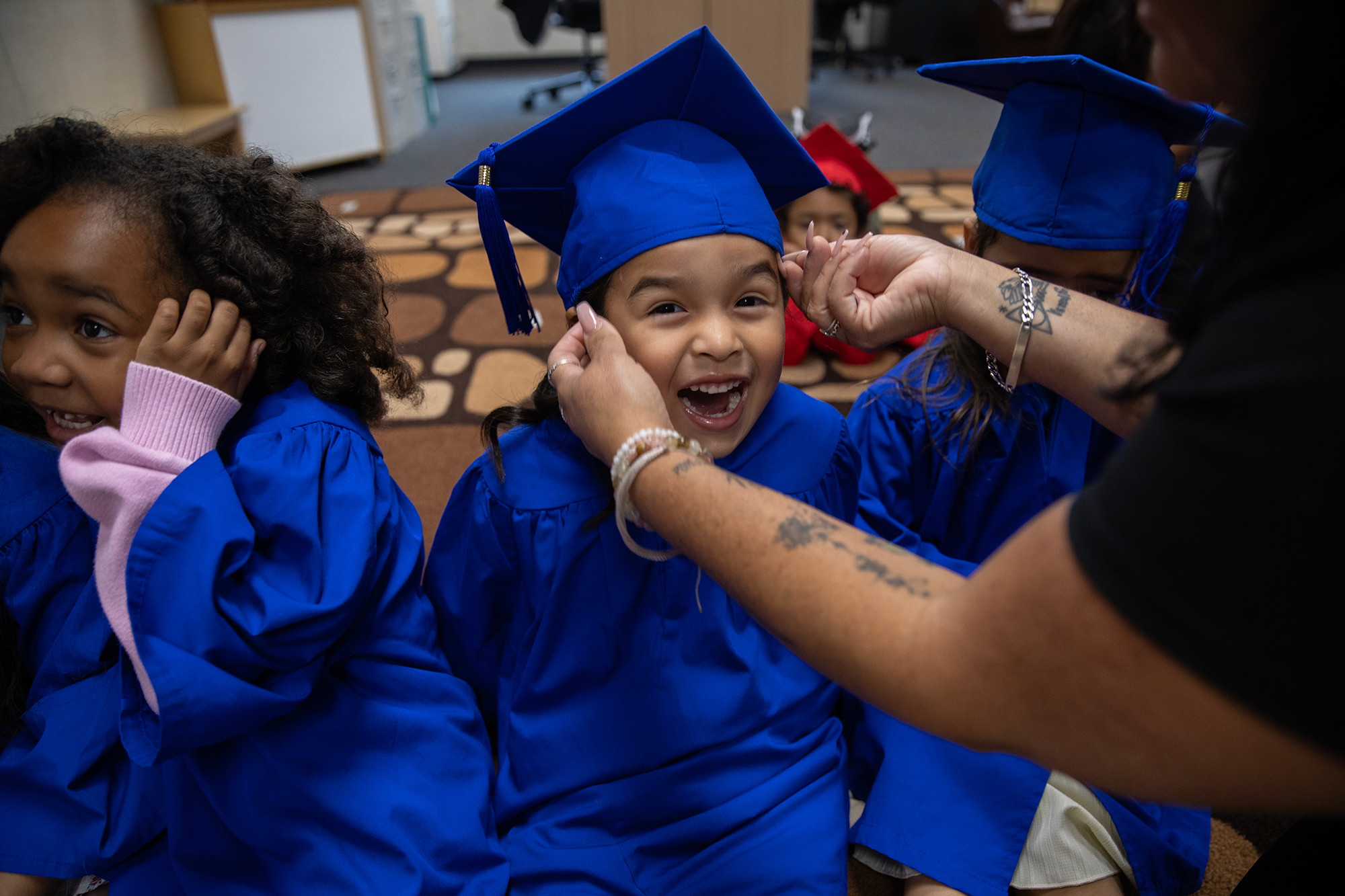 A child, wearing a blue graduation gown, smiles in excitement as an teacher adjusts a matching blue graduation cap on their head.
