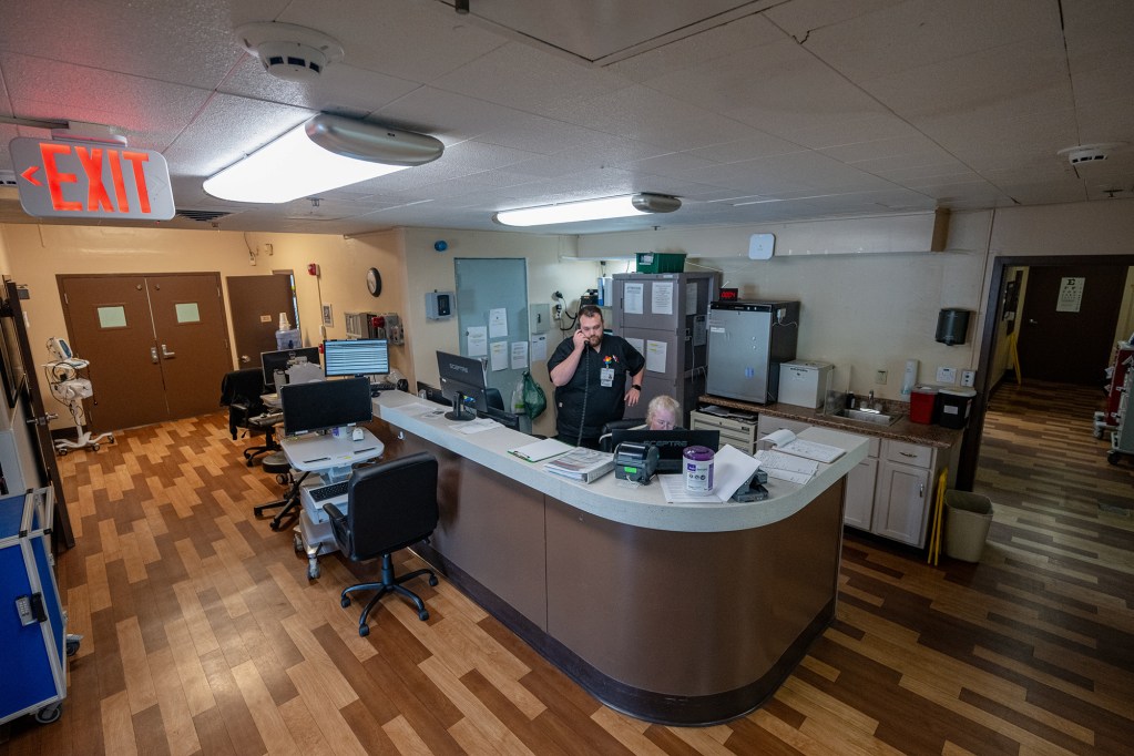 A wide view of a person on the phone as they stand behind a large rounded desk in the middle of a hospital building department.