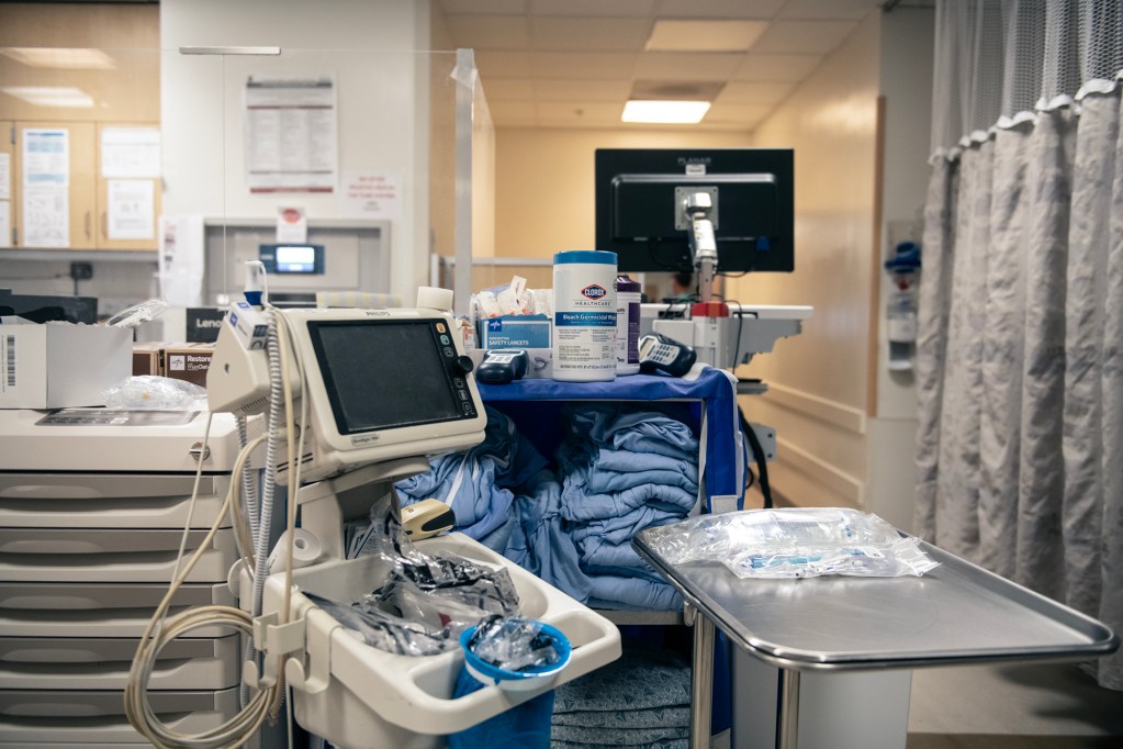 Medical equipment and supplies are organized on rolling carts in a hospital hallway. A monitor, disinfecting wipes, and folded blue gowns are visible, with a computer workstation in the background and privacy curtains along one side of the corridor.