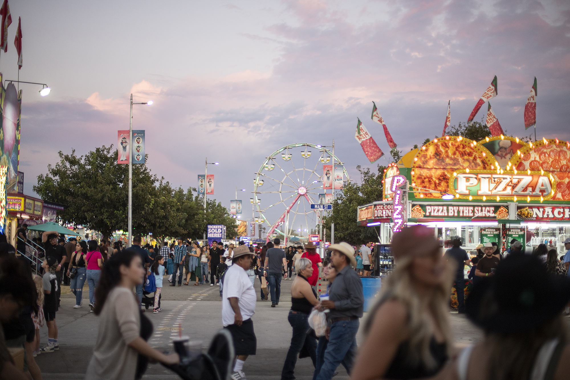 Various fairgoers walk through a fair group with a giant Ferris wheel in the background and multiple booths selling food. The photo depicts a busy scene of fairgoers enjoying various activities at a fair.