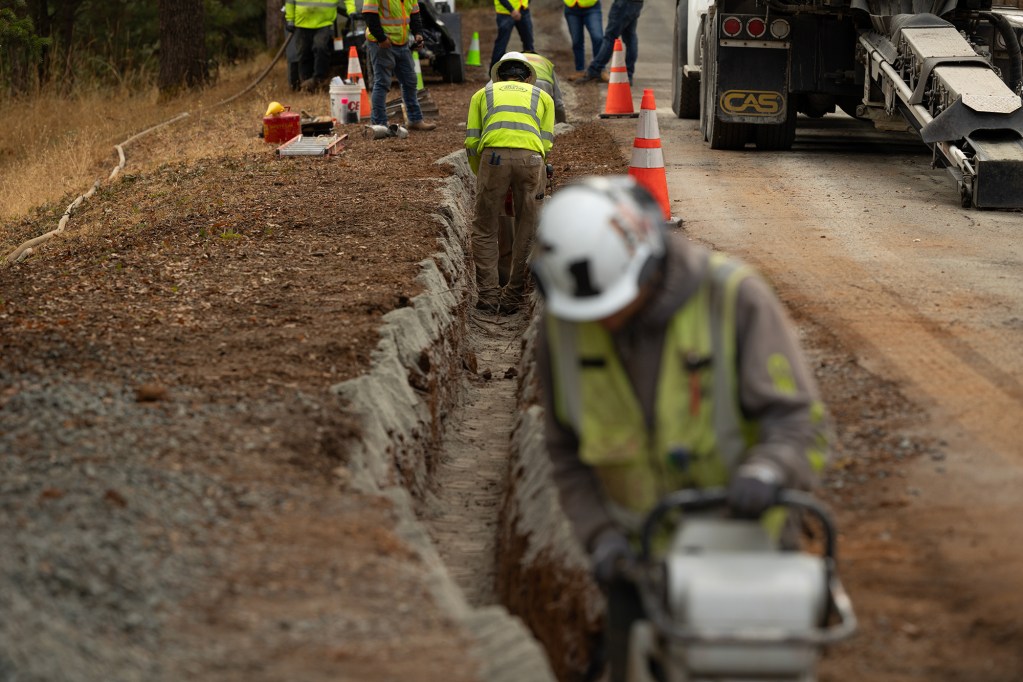 Construction workers use construction machinery as they work on a trench near the side of a road.