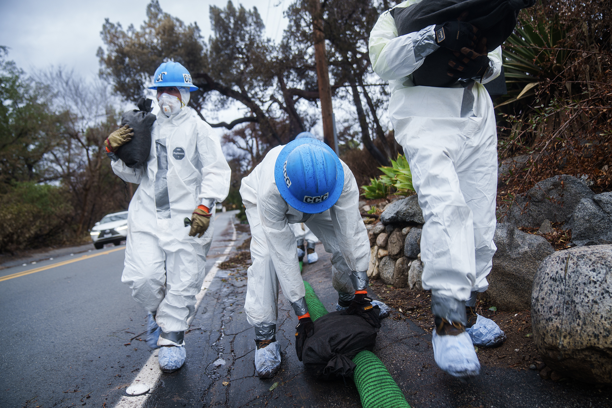 Workers in white protective suits and blue helmets place sandbags around a green hose along a wet roadside, with burned trees and debris visible in the background after severe weather and fire damage.