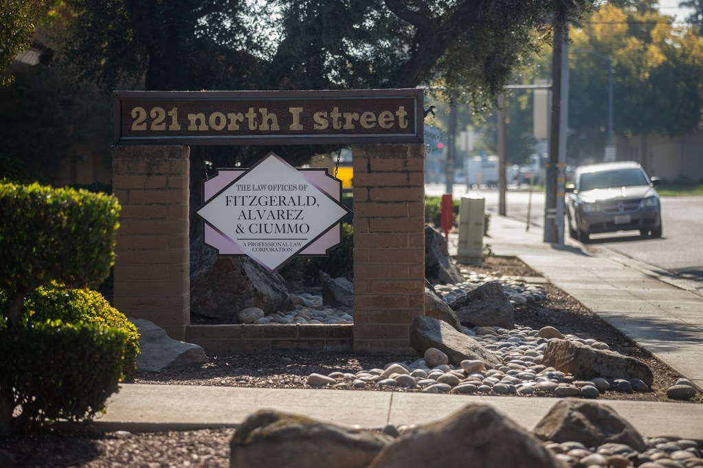 A brick sign at a street corner reads “221 North I Street” above a hanging plaque for “Fitzgerald, Alvarez & Ciummo, A Professional Law Corporation,” surrounded by rocks, shrubs, and a parked car in the background.
