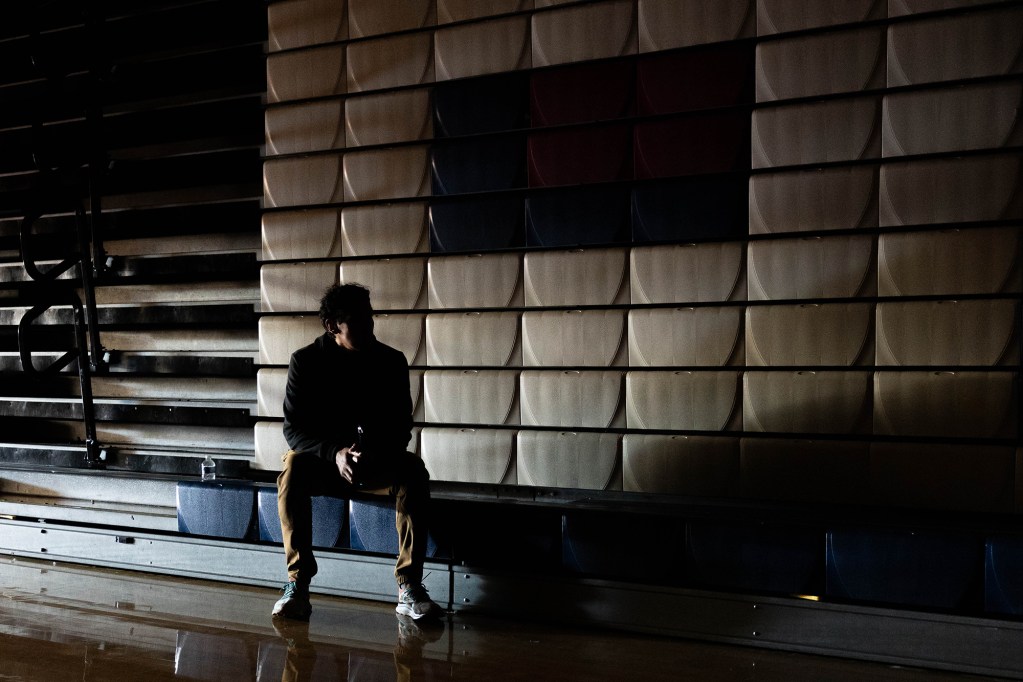 A person wearing a sweatsuit and sneakers is sitting on a set of bleachers in a dark gym, with light coming from one side of the room, creating a silhouette of the person and darkening their face to protect their identity.
