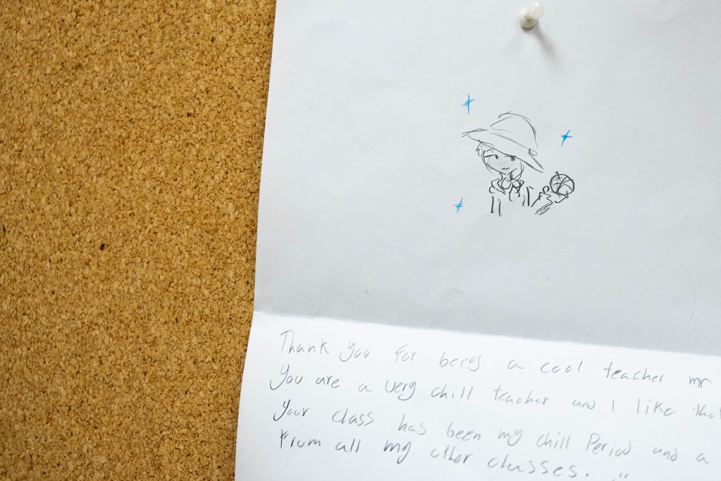 A piece of paper pinned to a corkboard with a thumb tack. The paper has a cartoon person drawn on one half, with a fold in the middle and a letter written on the bottom half.