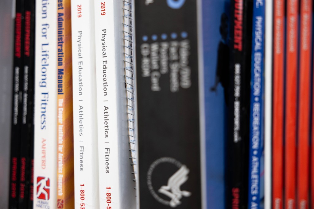 A close-up view of a row of books sitting on the shelf of a bookshelf, with the spines of two white books in focus, reading “Physical Education Athletic Fitness” on their spines.
