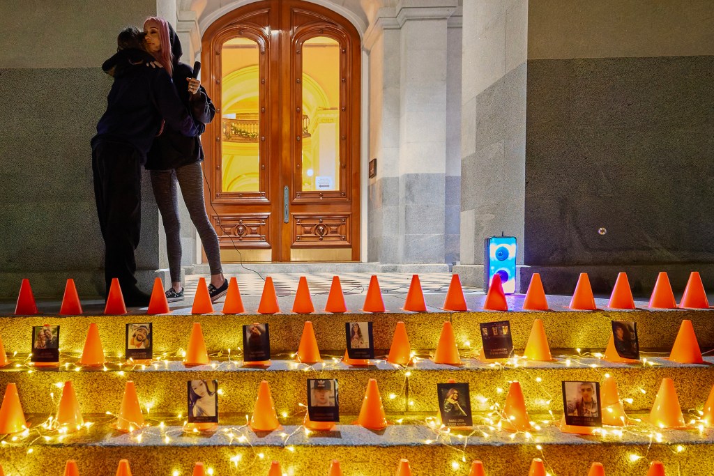 On the steps of a building at night, two people hug beside a microphone, while rows of small orange traffic cones and string lights line the stairs, each cone paired with a photo of a person, creating a memorial-like display.