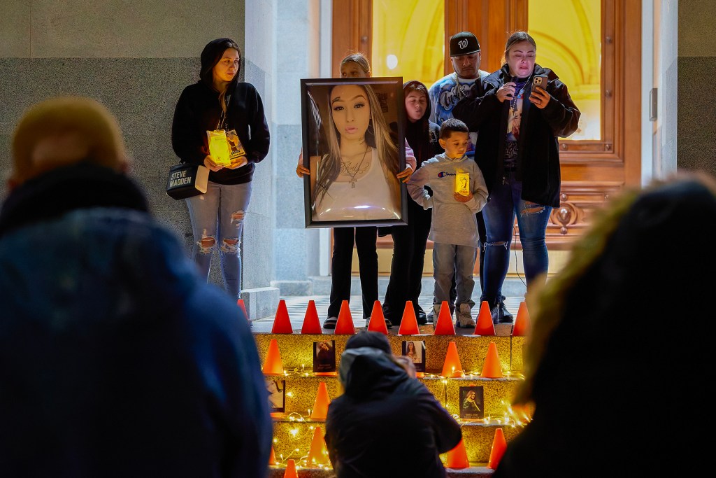 People stand on the steps of a building during a nighttime vigil, holding candles and a large framed portrait of a young woman. Orange cones and small memorial photos line the stairs, illuminated by string lights, while a speaker reads from a phone and others face the crowd.