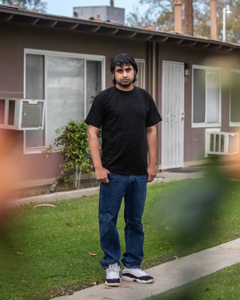 A person — with shaggy black hair and wearing a black shirt with blue jeans — stands in a cement path in front of two houses.