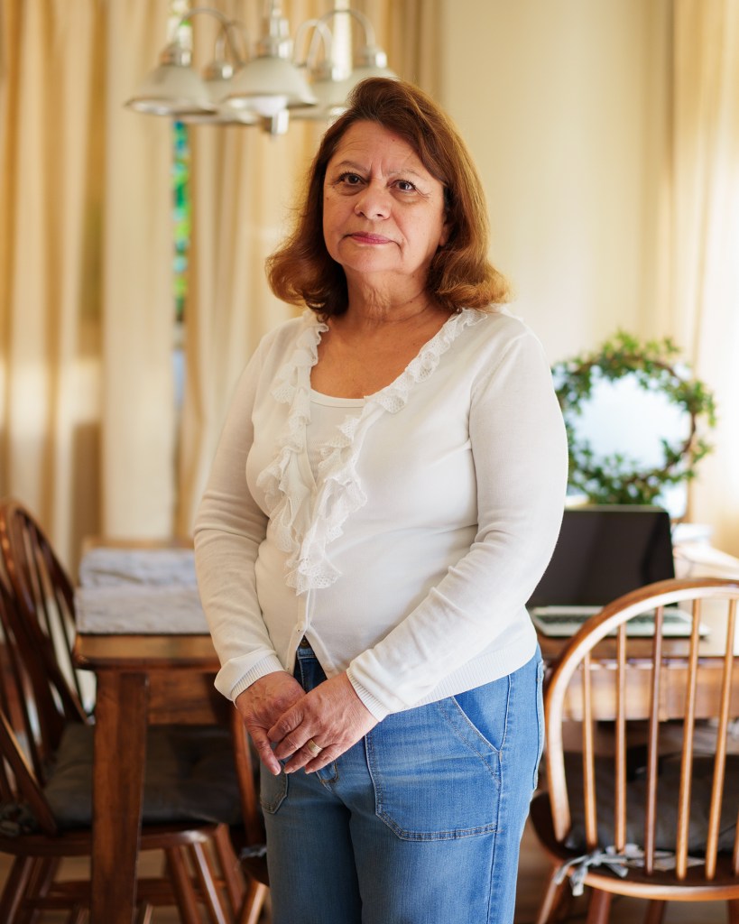 A person — with red short hair and wearing a white blouse and blue jeans — stands in front of a dining table inside a house.