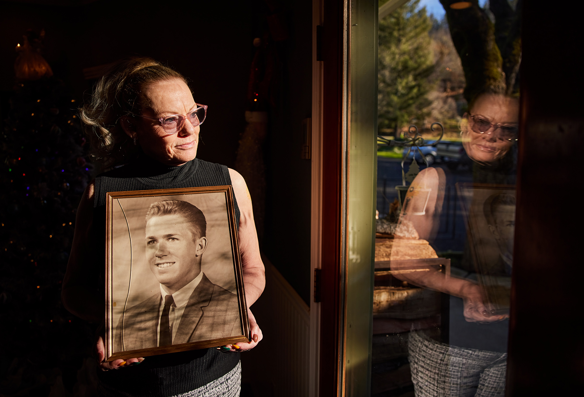 A person in glasses holds a wooden-framed black-and-white photograph to her chest while standing by a window in a dark room. The woman is looking outside of the window while she and the picture are reflected on the glass window.