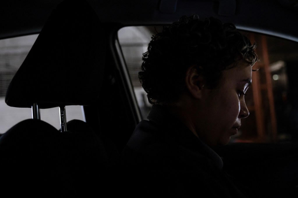 A person sits in the front seat of a car, shown in profile, with short curly hair softly lit by dim light from outside the vehicle. The interior is mostly dark, with the headrest, window frame, and a blurred structure visible beyond the window, creating a quiet, contemplative mood.