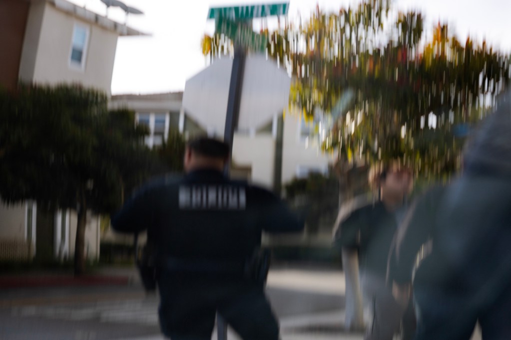 A motion-blurred street scene shows a uniformed officer moving quickly near an intersection, with another person partially visible nearby. A stop sign and street sign appear in the background alongside residential buildings and trees, conveying a sense of sudden movement and urgency.