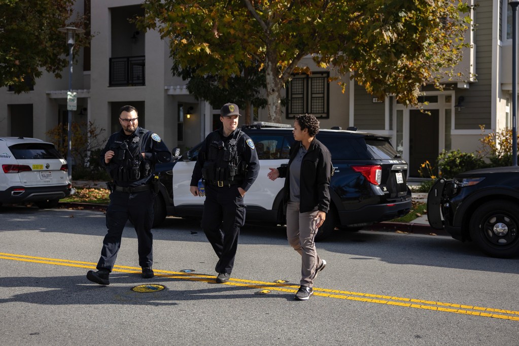 Three people walk together across a residential street, including two uniformed police officers and a third person in plain clothes who gestures while speaking. Parked police vehicles line the curb behind them, and apartment buildings and trees with autumn leaves frame the scene in daylight.